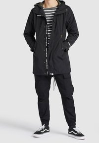 Personne portant une veste à capuche noire, un pantalon cargo noir, des baskets noires et blanches, et une chemise rayée gris et blanc, les mains dans les poches.