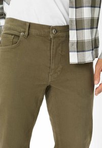 Pantaloni in cotone verde oliva con una vestibilità slim. Presentano una chiusura a bottone, tasche laterali e dettagli di cucitura sottili. Abbinati a una camicia a quadri.