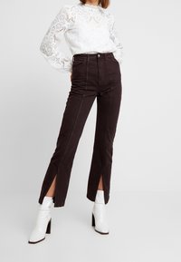 Personne portant un pantalon taille haute marron foncé avec des fentes à l'avant, associé à des bottines blanches à talons et un haut en dentelle blanche à manches longues.