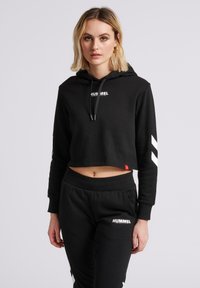 Blondynka w czarnej, skróconej bluzie z kapturem i pasujących czarnych spodniach dresowych z białymi logo "Hummel", stojąca na jednolitym jasnym tle.