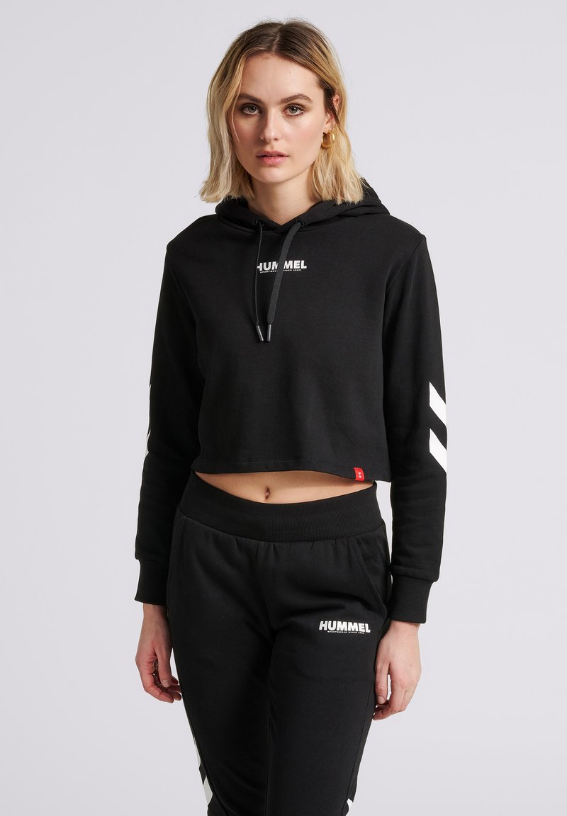 Blondynka w czarnej, skróconej bluzie z kapturem i pasujących czarnych spodniach dresowych z białymi logo "Hummel", stojąca na jednolitym jasnym tle.