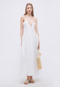 Žena v bielych maxi šatách bez rukávov so spaghetti ramienkami, drží pletenú slamovú tašku a má na nohách béžové šľapky.