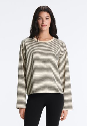 Frau trägt ein beiges Langarmshirt mit dünnen horizontalen schwarzen Streifen und schwarze eng anliegende Hosen, steht vor einem weißen Hintergrund.