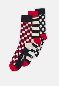 Trois paires de chaussettes : une avec des motifs géométriques rouges et noirs, une avec des rayures rouges et blanches, et une avec des pois blancs sur fond noir.