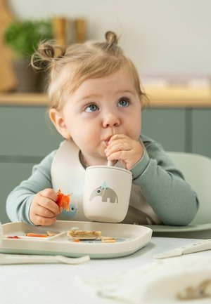 Dieťa drží mäkký, béžový pohár s motívom slona, má na sebe svetlomodré tričko s dlhým rukávom a hrá sa s jedlom na rozdelenej tácke.