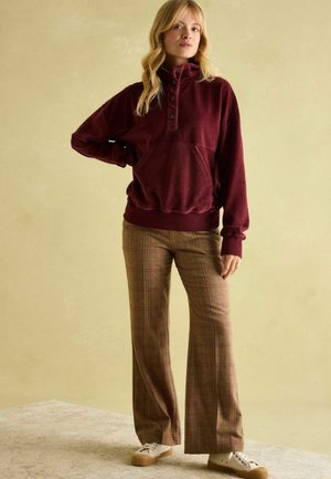 Burgundy corduroy pullover with a high collar, button details, and a front pocket, paired with brown checked wide-leg trousers and beige sneakers.