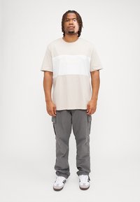 Beige t-shirt met korte mouwen en een witte horizontale strook met reliëftekst, gecombineerd met grijze cargobroek en witte sneakers.