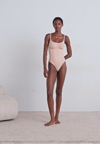 Nude fitted bodysuit with thin straps, smooth texture, high-cut legs, and seamless design. Model stands on a light carpet against a pale wall.