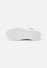 Valge sneaker'i tallaga, millel on tekstureeritud ja soonikkoes muster, musta aksendi ja graveeritud logoga. Sile pind ja ümarad servad.