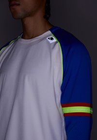 Sportowa koszulka z długim rękawem, z beżowym przodem, niebieskimi, teksturowanymi rękawami oraz żywymi zielonymi, czerwonymi i niebieskimi akcentami na ramionach.