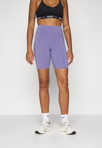 Shorts de vélo taille haute violets avec une texture lisse, associés à un brassière de sport noire avec une bande logo blanche et des baskets de couleur claire.