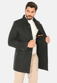 Cappotto sartoriale grigio scuro con colletto alto, chiusura con bottoni e interno trapuntato con dettaglio tasca arancione. Tessuto dalla trama liscia.
