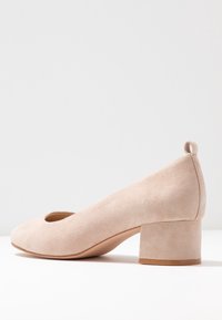 Chaussure femme beige en daim avec petit talon bloc, bout arrondi et languette à l'arrière, présentée sur une surface blanche et un fond clair.