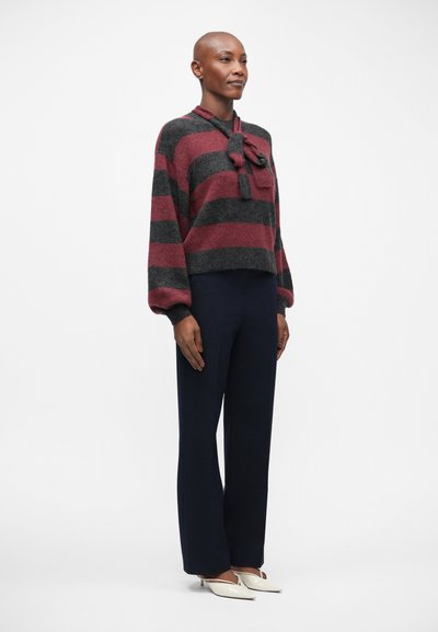 Pull rayé dans des tons de rouge foncé et gris, avec un détail nœud au cou. Associé à un pantalon marine et des chaussures blanches à bout pointu.