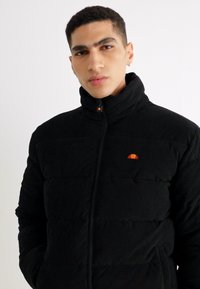 Schwarze Cord-Puffermantel mit hohem Kragen, versehen mit einem kleinen orange- und gelbfarbenen Logo auf der linken Brust und mit Reißverschlusstaschen.