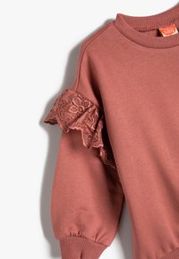 Różowy sweter z falbanami wykonany z delikatnego materiału, z koronkowymi akcentami na ramionach, ściągaczami przy mankietach i dekolcie; miękka tekstura i swobodny krój.