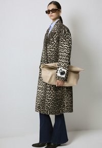 Femme portant un manteau en imprimé léopard, une chemise bleue, un jean évasé, des bottes noires, des lunettes de soleil en écaille de tortue, tenant un grand sac à main beige.