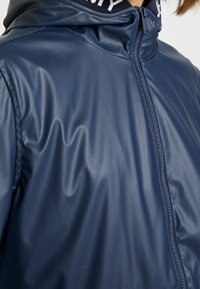 Laivansininen vedenpitävä takki, joka on sileä ja kiiltävä pinta; siinä on vetskari edessä ja huppu, jossa on kontrastiväri kauluksessa.