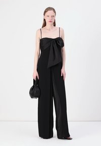 Combinaison noire avec un grand nœud au niveau de la poitrine, bretelles spaghetti, jambes larges et finition satinée lisse. Porte un petit sac à main noir.