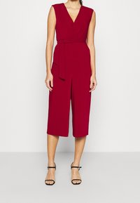 Combinaison sans manches rouge avec un décolleté en V, taille cintrée, design enveloppant et jambes larges coupées. Associée à des sandales noires à brides.