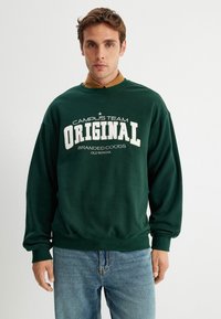 Groene sweatshirt met witte tekst "CAMPUS TEAM ORIGINAL" en "BRANDED COODS OLD SCHOOL." Katoenen materiaal. Relaxed fit en ribgebreide manchetten. Draagt met blauwe spijkerbroek.