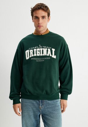 Grüner Pullover mit weißem Text "CAMPUS TEAM ORIGINAL" und "BRANDED COODS OLD SCHOOL." Aus Baumwollmaterial. Lockere Passform und gerippte Bündchen. Getragen mit blauen Jeans.