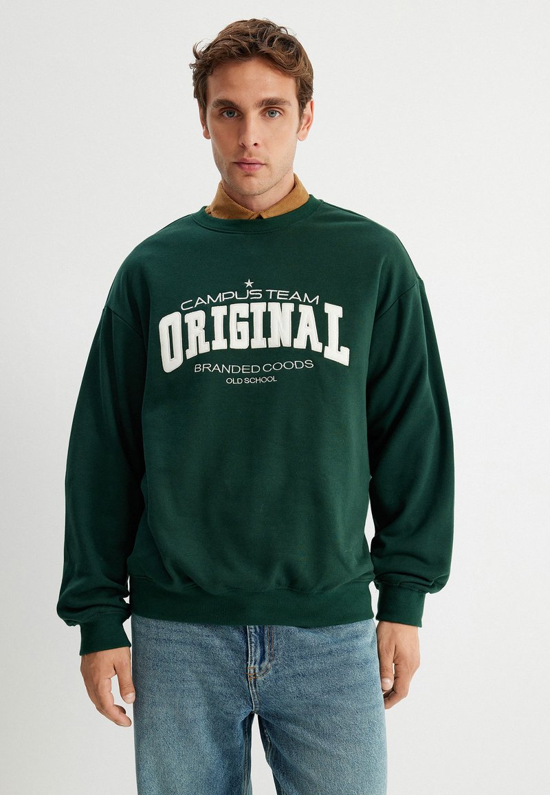 Sudadera verde con texto blanco "CAMPUS TEAM ORIGINAL" y "BRANDED COODS OLD SCHOOL". Material de algodón. Corte relajado y puños acanalados. Llevada con pantalones vaqueros azules.