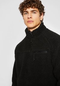 Sort fleece pullover med høj krave, halv-lynlås foran, tekstureret overflade og en lynlåslommes til brystet med sorte hardware-detaljer.