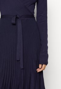 Femme portant une robe en maille côtelée bleu marine avec une ceinture nouée à la taille et des manches longues, la main posée près de la jupe.