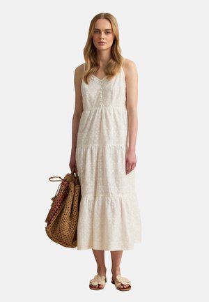 Frau mit langen blonden Haaren, die ein ärmelloses weiß besticktes Midikleid trägt, eine große geflochtene braune Tragetasche hält und beige Slide-Sandalen trägt.