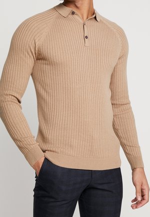 Mężczyzna w beżowym prążkowanym swetrze polo z długim rękawem i dwoma guzikami, z rękami w kieszeniach ciemnych, kraciastych spodni, na jednolitym tle.