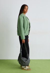 Casaco verde puxado, calças pretas, saco cinza xadrez e sapatilhas verdes com riscas brancas. Cabelo trançado, em pé sobre um chão verde.