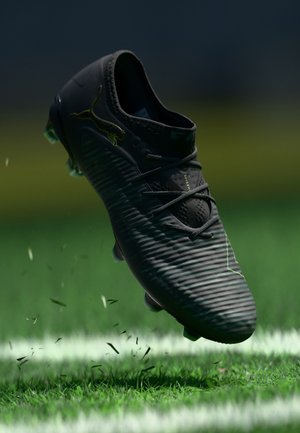 Sort Puma fodboldstøvle med snørebånd midt i luften over grøn græsmark med hvid linjemarkering, små græsstykker flyver rundt om sålen.