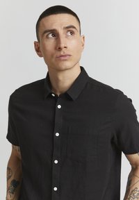 Sort kortærmet skjorte med knapper, lavet af tekstureret stof, med en brystlomme og hvide knapper langs knaplisten.