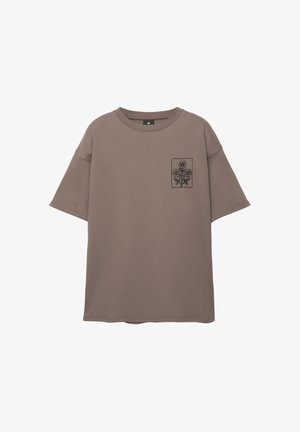 T-shirt marrone oversize realizzato in morbido cotone, con un piccolo ricamo floreale nero nella zona del taschino sinistro.