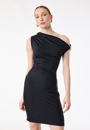 Nainen, joka pitää päällään vartalonmyötäistä mustaa yksisojaista mekkoa, jossa on rypytettyjä yksityiskohtia, sekä kultaiset renkorenkaat korvakoruissa, seisoo yksinkertaisen valkoisen taustan edessä.