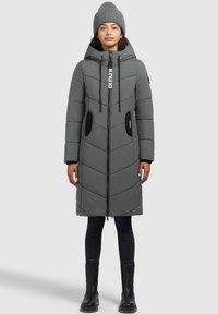 Donna che indossa un cappotto imbottito grigio fino al ginocchio con cappuccio, zip nera, dettagli neri sulle tasche, berretto a maglia grigio, pantaloni neri e stivali neri.