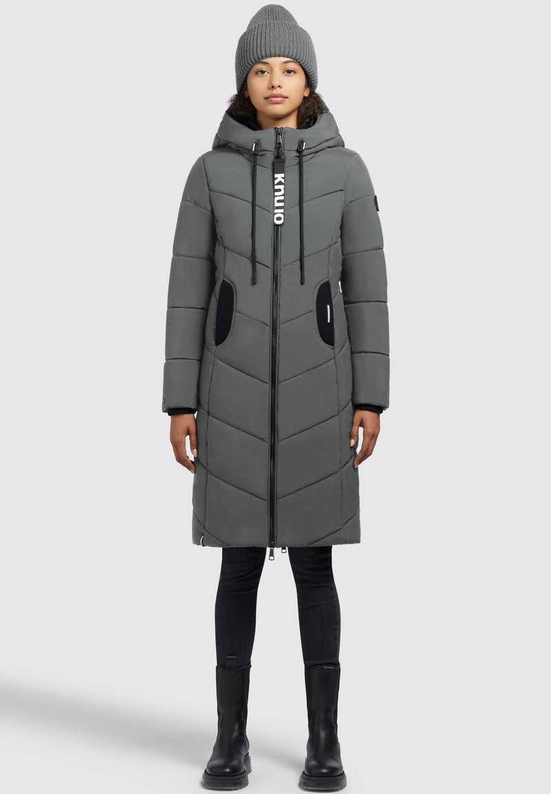 Donna che indossa un cappotto imbottito grigio fino al ginocchio con cappuccio, zip nera, dettagli neri sulle tasche, berretto a maglia grigio, pantaloni neri e stivali neri.