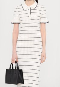 Robe blanche à rayures horizontales noires, manches courtes à volants et col. Tenant un petit sac à main en cuir noir avec des poignées.