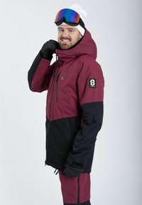 Maroonskijakke med sort, justerbar hætte, lynlåslommer og logo-patch på ærmet; bæres med farverige goggles og handsker.