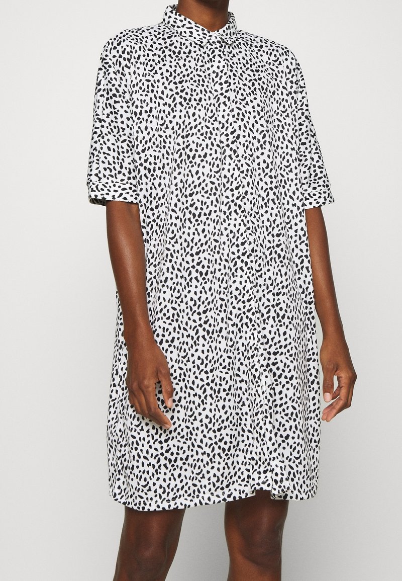Robe à manches courtes, longueur genoux en tissu blanc avec un motif de taches noires. Présente un devant à boutons et un col.