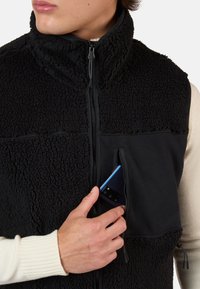 Gilet in pile nero con una superficie testurizzata e pannelli lisci. Presenta una tasca con zip per un telefono. La mano del modello è visibile mentre si avvicina alla tasca.