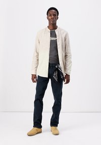Camisa de lino beige de botón sobre una camiseta gris, jeans azul oscuro y zapatillas beige. Bandana negra atada a la cintura.