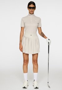 Kobieta ubrana w beżowy strój golfowy, okulary przeciwsłoneczne i rękawiczkę, trzymająca kij golfowy, stojąca na jednolitym tle.