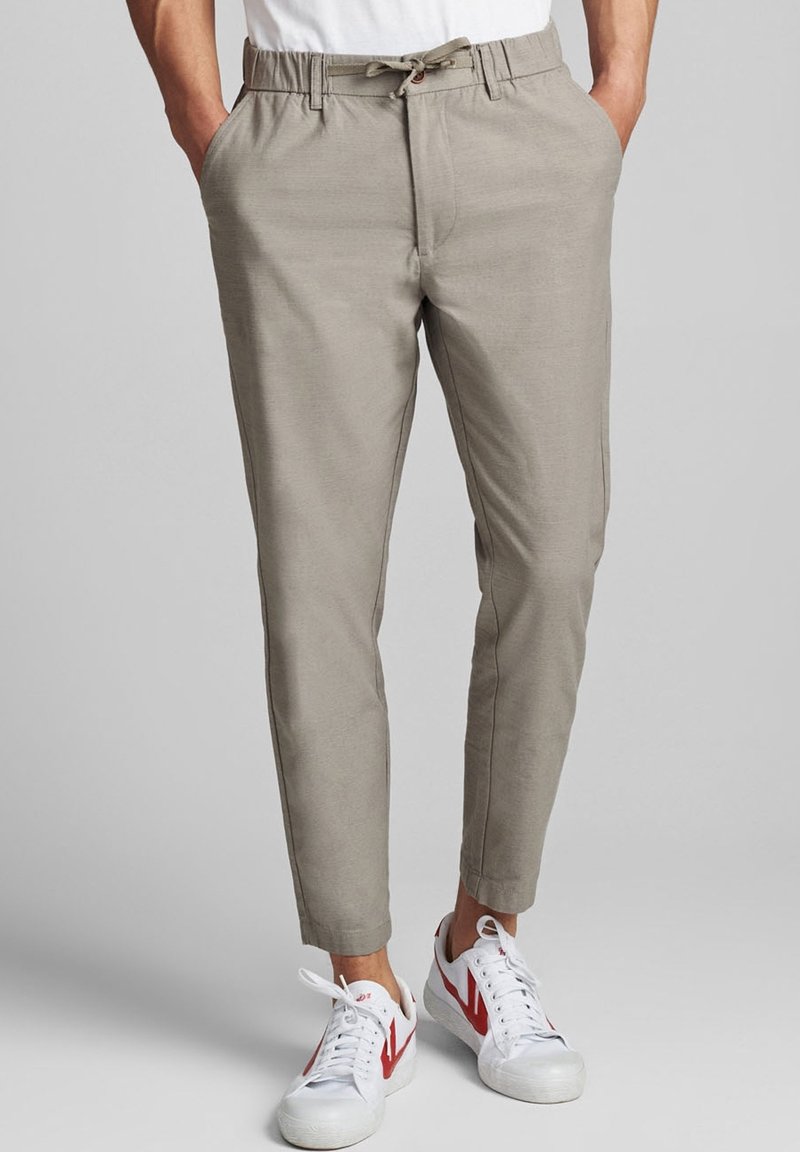 Beżowe spodnie o luźnym kroju, z elastycznym paskiem na sznurku i kieszeniami, w połączeniu z białymi sneakersami z czerwonymi akcentami. Gładka struktura materiału.