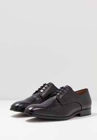 Une paire de chaussures de ville en cuir noir poli avec lacets sur une surface blanche, le tout sur un fond gris clair uni.