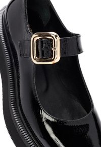 Chaussure Mary Jane en cuir verni noir avec une bride à boucle dorée, un bout arrondi et une semelle en caoutchouc noire texturée. Finition intérieure lisse.