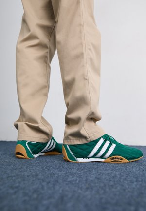 Zapatillas de deporte verdes acolchadas con rayas blancas y suelas de goma marrón, combinadas con pantalones beige. El diseño presenta un perfil bajo y material texturizado.
