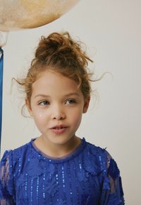 Abito blu realizzato in tessuto trasparente con applicazioni floreali e decorazioni in paillettes. Bambino con capelli ricci sta in piedi accanto a un palloncino.
