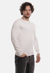 Maglione bianco a maglia con colletto tondo, design aderente e tessuto strutturato. Il modello indossa jeans scuri, con le mani in tasca, in piedi su uno sfondo bianco.
